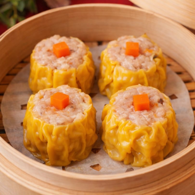 Four steamed Siu Mai dumplings with orange carrot garnishes in a bamboo steamer basket.
