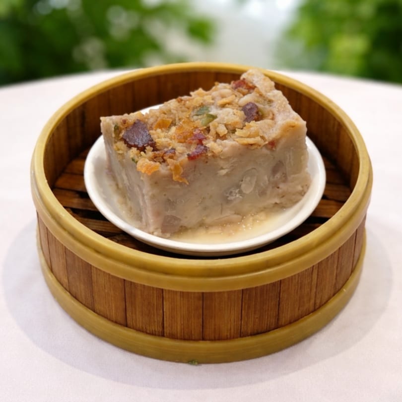 A slice of taro cake with crispy toppings, served in a bamboo steamer on a white plate.